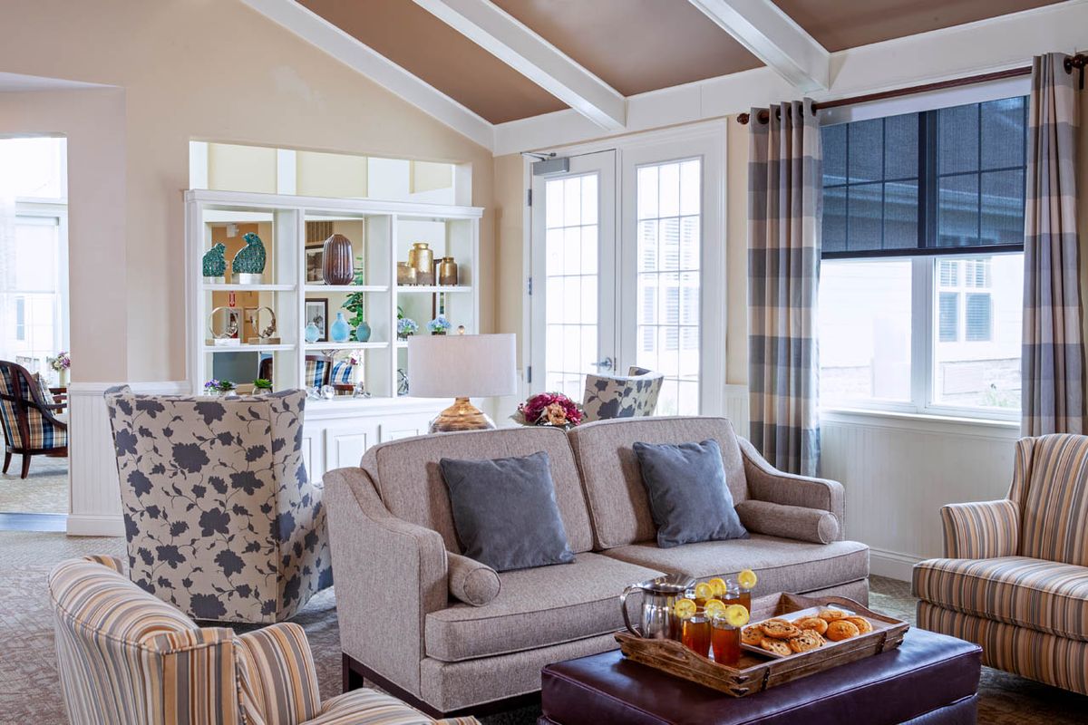 A cozy living room featuring a comfortable sofa with cushions, a coffee table with refreshments, and elegant decor set against large windows and decorative shelves.