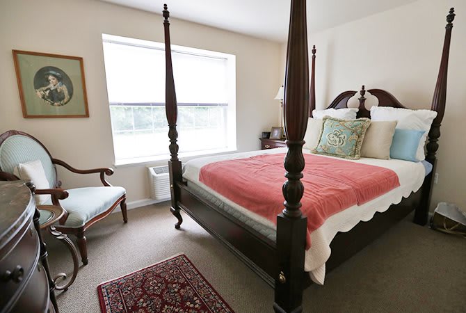 A cozy bedroom featuring a four-poster bed with a red blanket, an upholstered chair, and a window providing natural light.