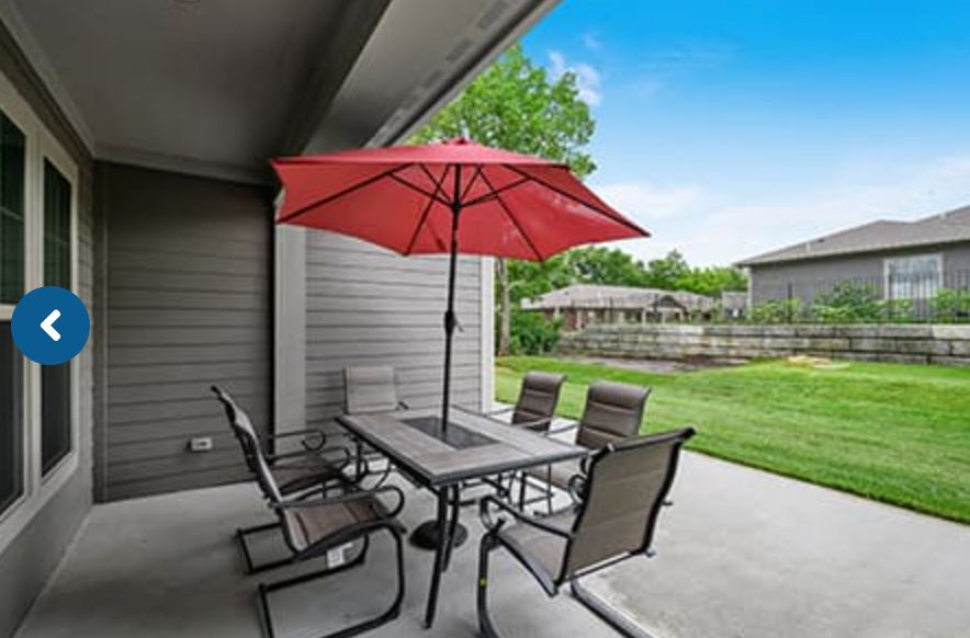 An outdoor patio setup with a table, chairs, and a red umbrella in a residential backyard.