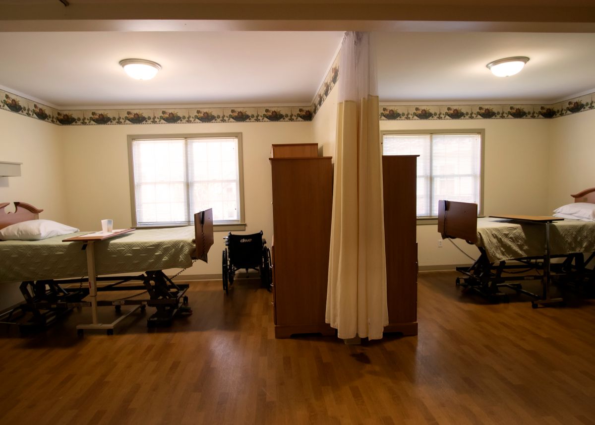 A shared room in a nursing home with two hospital beds, separated by a privacy curtain, and a wheelchair near the window.