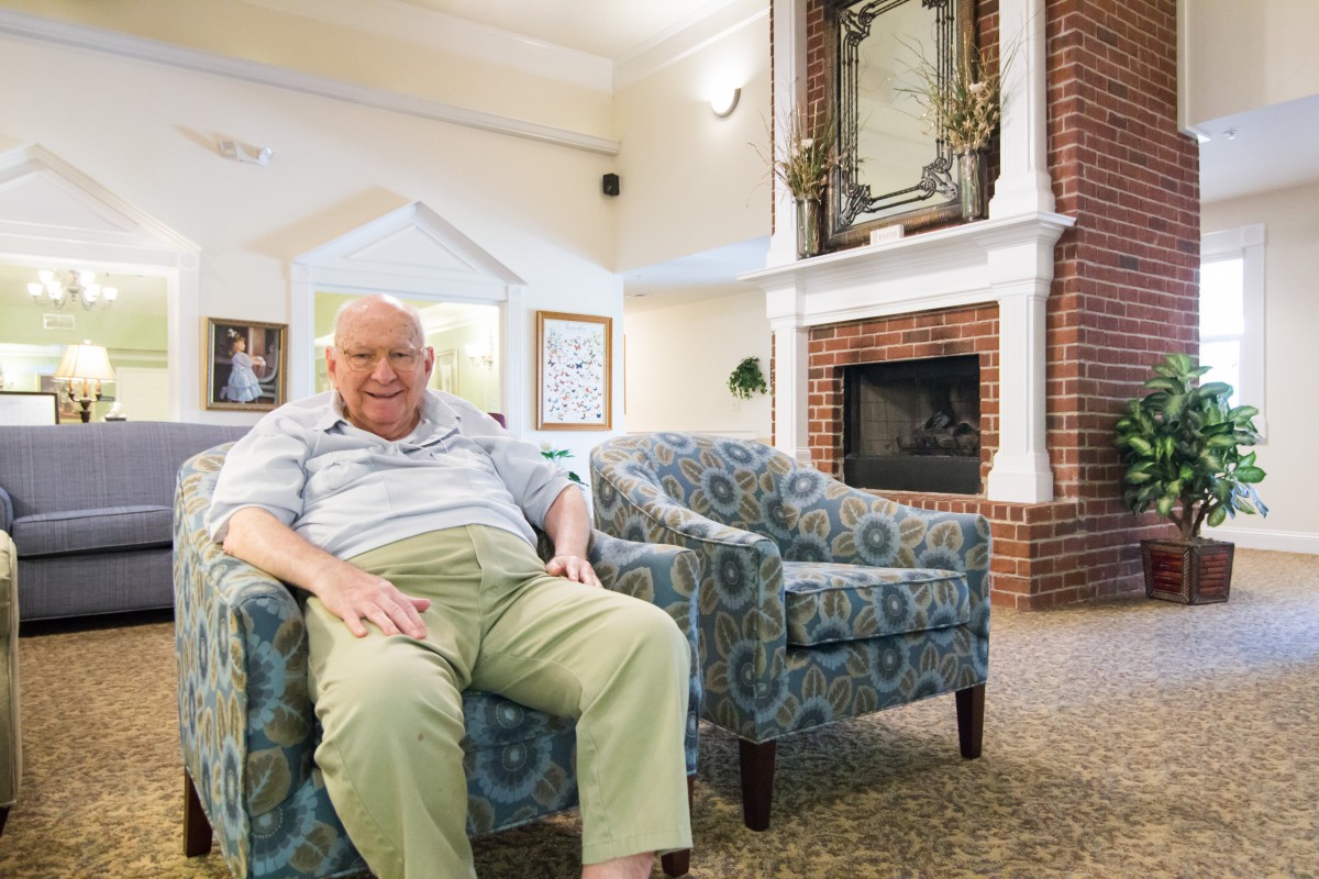 An elderly man sitting comfortably in a living room with cozy furniture and a fireplace.