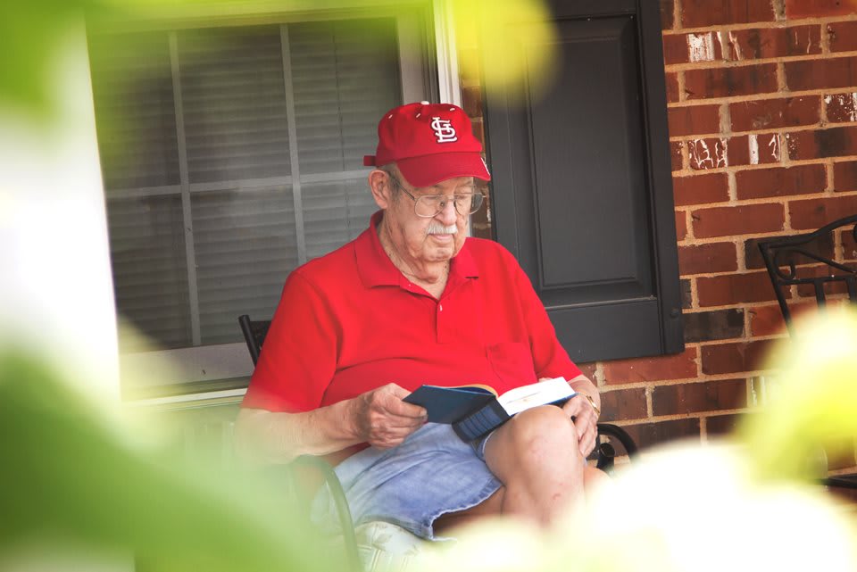An elderly man in a red shirt and cap sitting on a porch, reading a book.