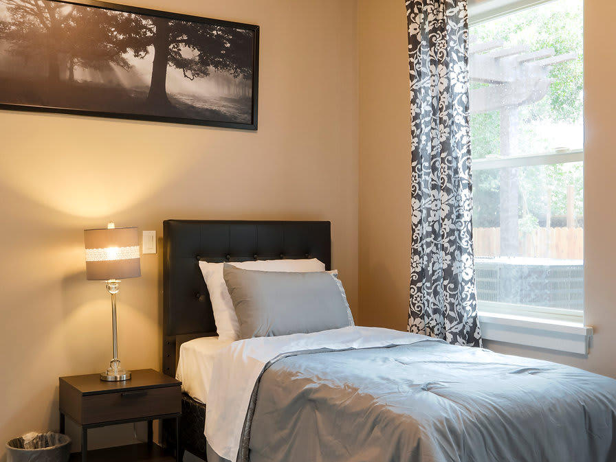 A cozy bedroom with a neatly made bed, a bedside lamp, and a window with patterned curtains.
