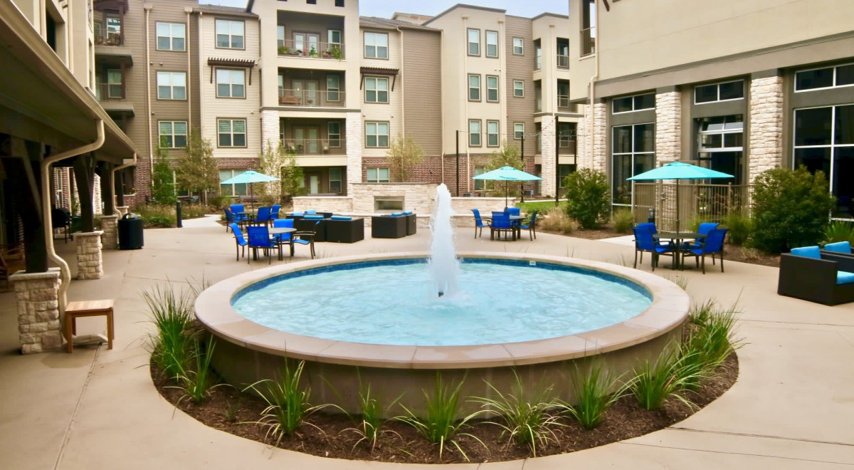 A modern apartment complex courtyard featuring a central fountain and outdoor seating with patio umbrellas.