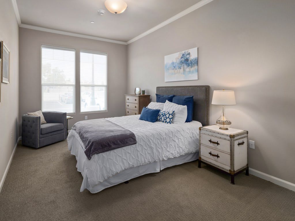 A cozy bedroom with a neatly made bed, nightstand with a lamp, and a window providing natural light.