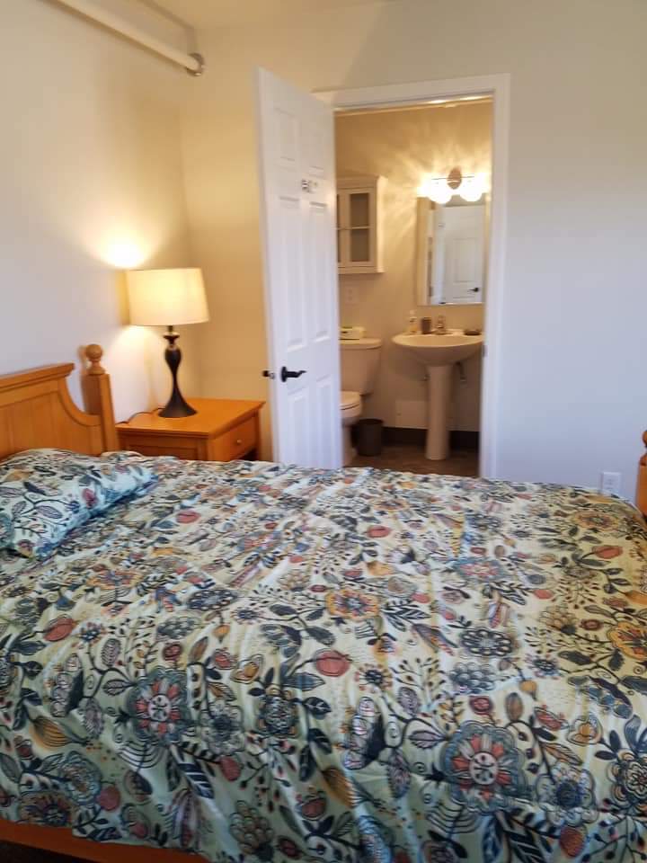 A cozy bedroom with a floral-patterned bedspread, wooden nightstand, and a view into an adjacent bathroom.