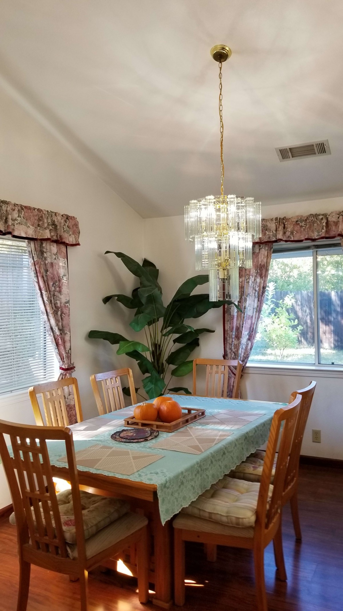 A cozy dining room with a table and chairs, adorned with a chandelier, indoor plant, and large windows with curtains.