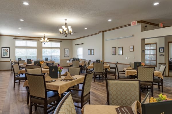 A cozy dining room featuring neatly arranged tables and chairs, illuminated by chandeliers.
