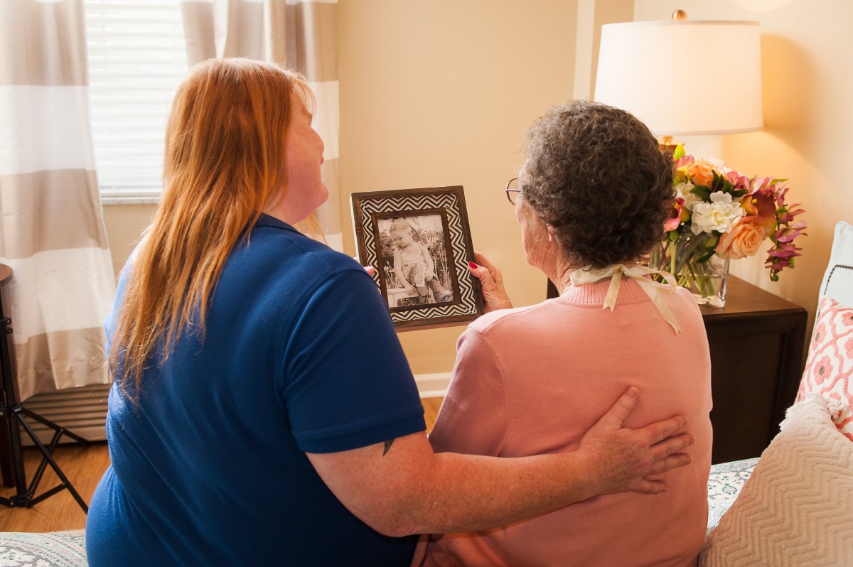 A caregiver and an elderly woman share a moment looking at a framed photograph.