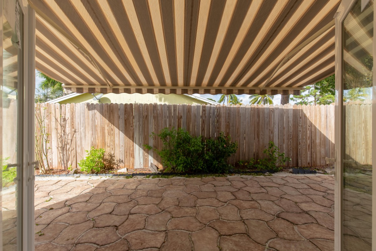 A patio with a striped awning providing shade, bordered by a wooden fence and stone flooring.