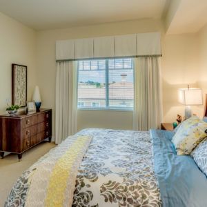 A cozy and well-decorated bedroom featuring a large bed with floral bedding, a wooden dresser, and a window with curtains.