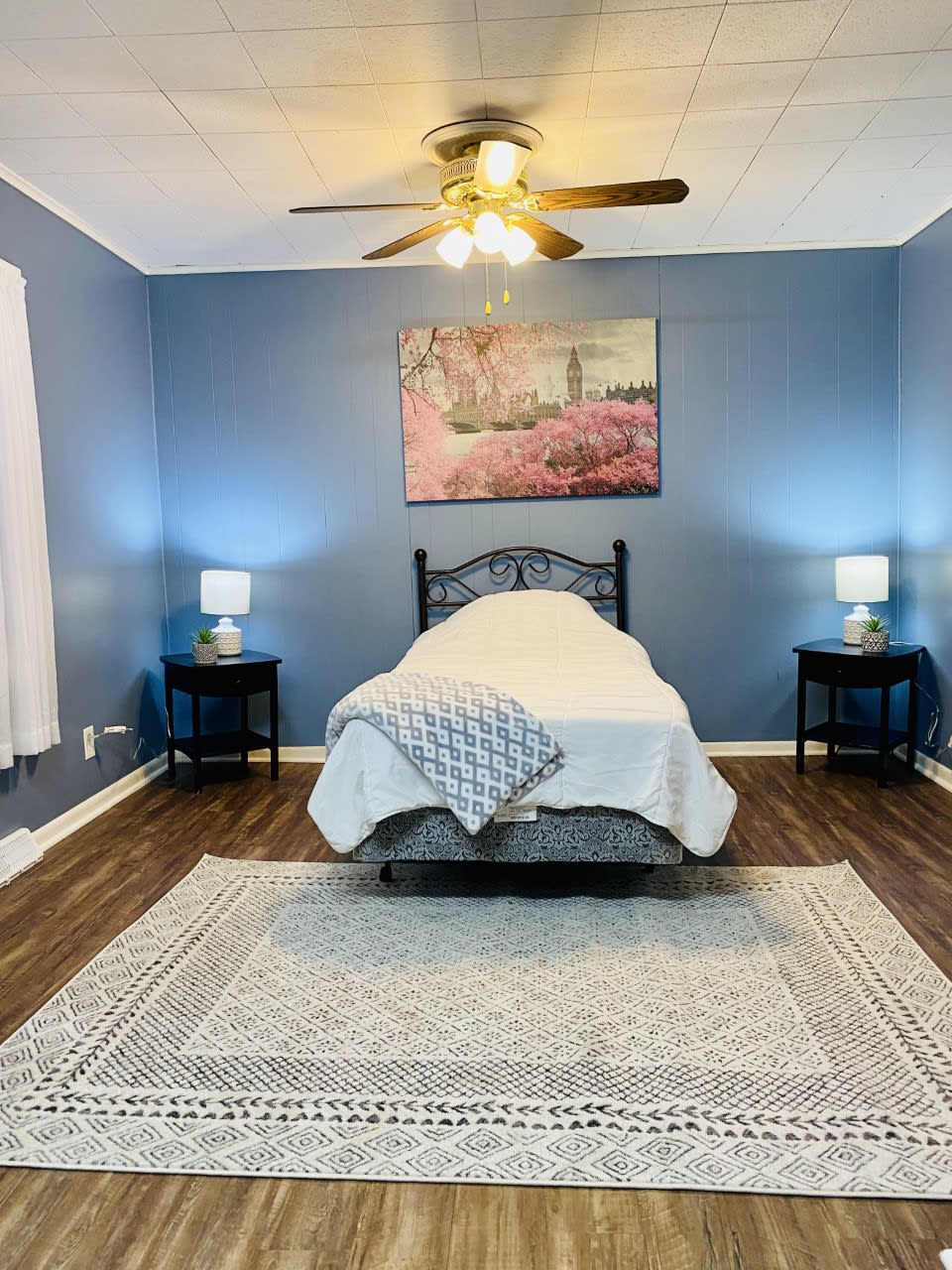 A cozy bedroom with blue walls featuring a single bed, nightstands with lamps, and a decorative rug.