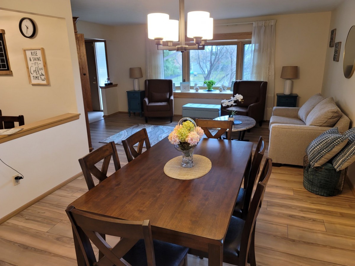 A cozy dining and living room area with a wooden table, chairs, and natural lighting from large windows.