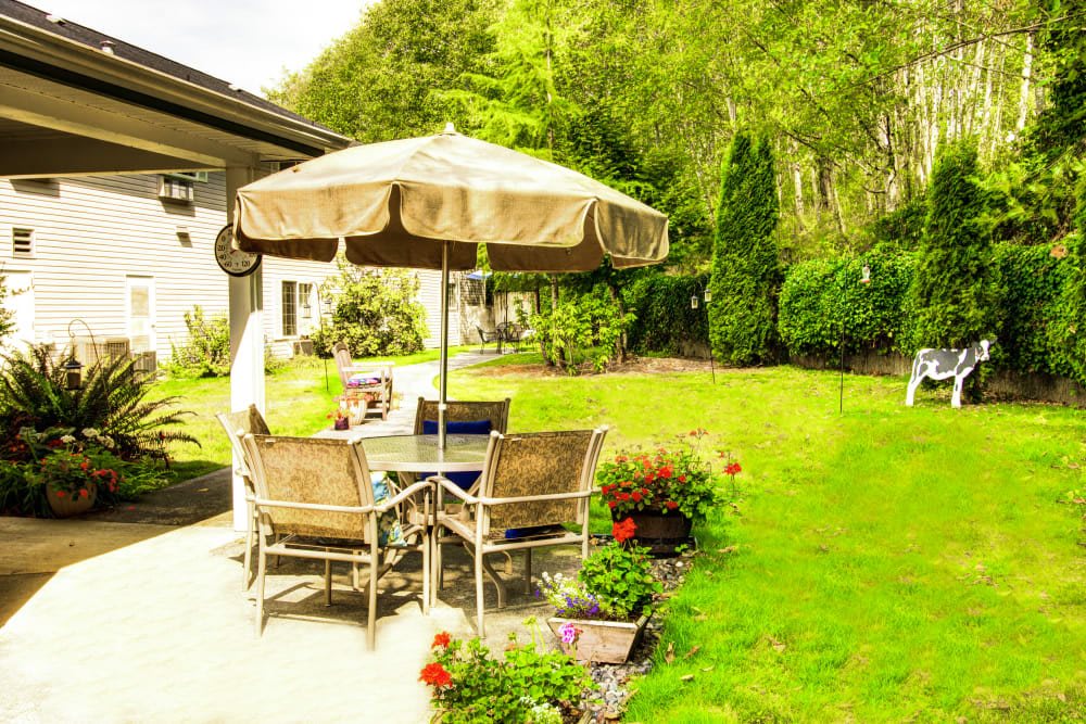 A sunny backyard patio with an umbrella and outdoor dining set surrounded by a lush green garden.