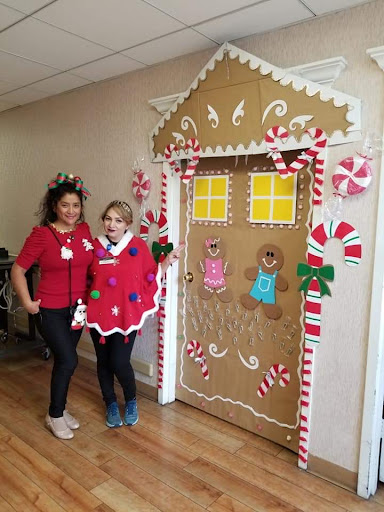 Two individuals in festive costumes pose next to a creatively decorated door resembling a gingerbread house, celebrating the holiday spirit.