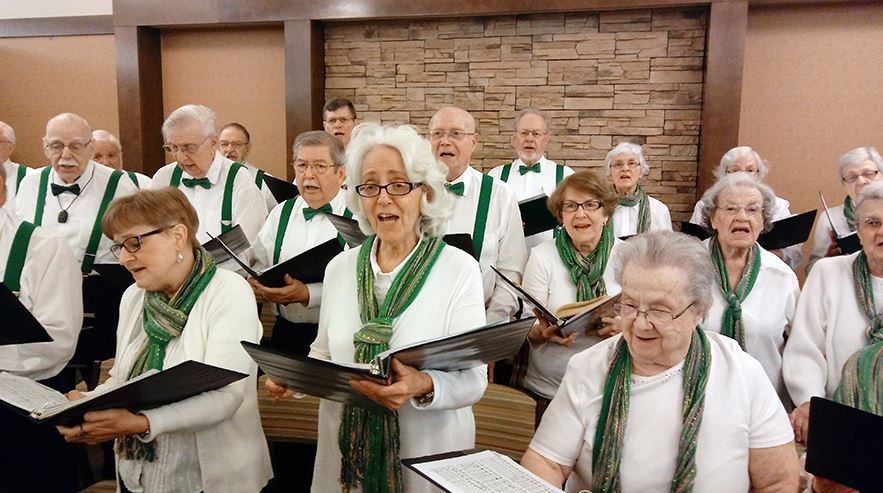 An elderly choir dressed in matching outfits performs together, holding sheet music.