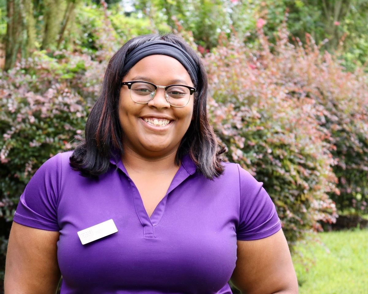 A woman in a purple shirt smiling outdoors with greenery in the background.