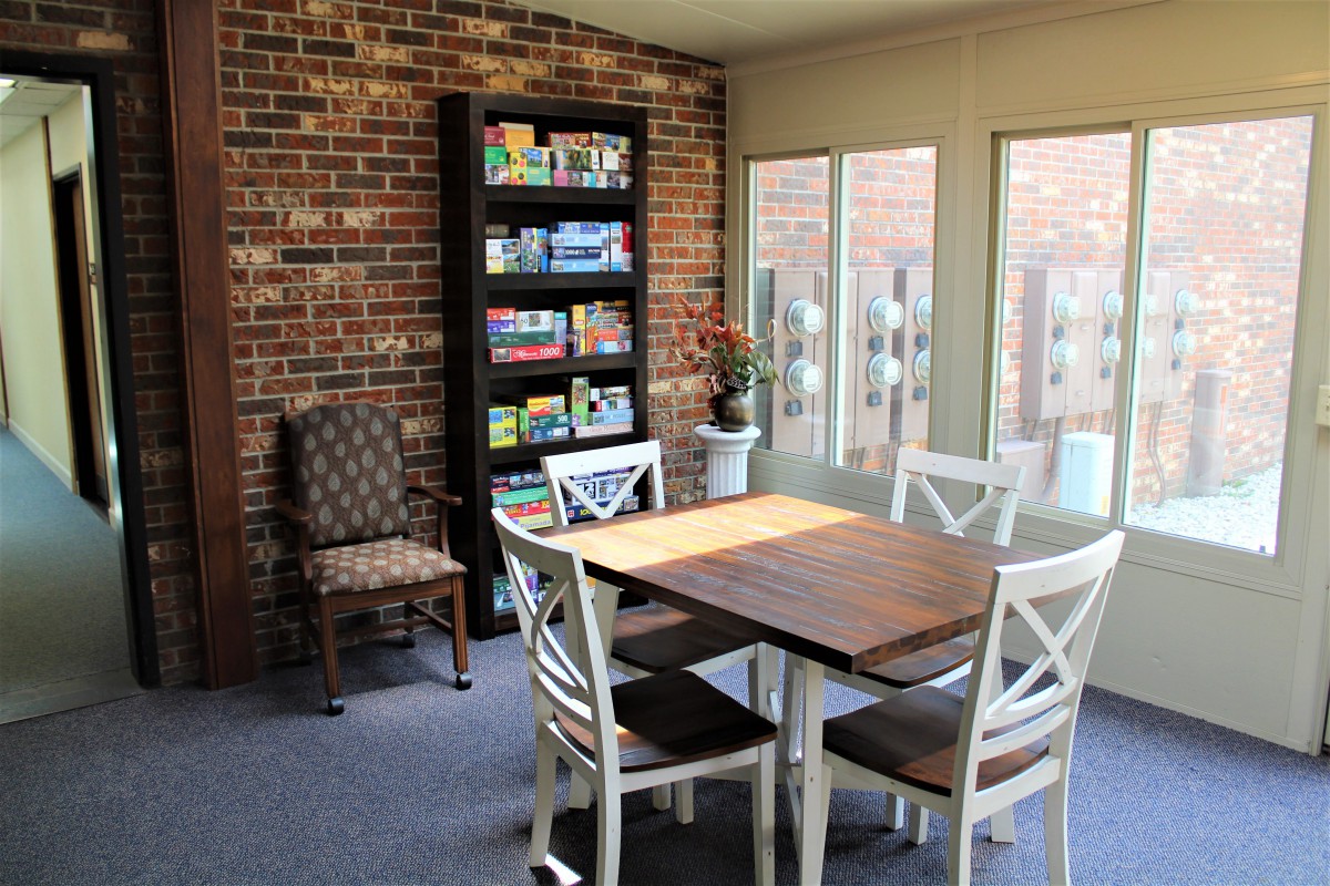 A cozy dining area with a wooden table, chairs, and a bookshelf against a brick wall, illuminated by natural light from large windows.
