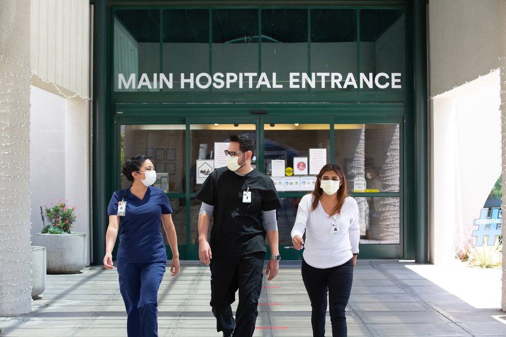 Healthcare workers wearing masks walk together at the main hospital entrance.