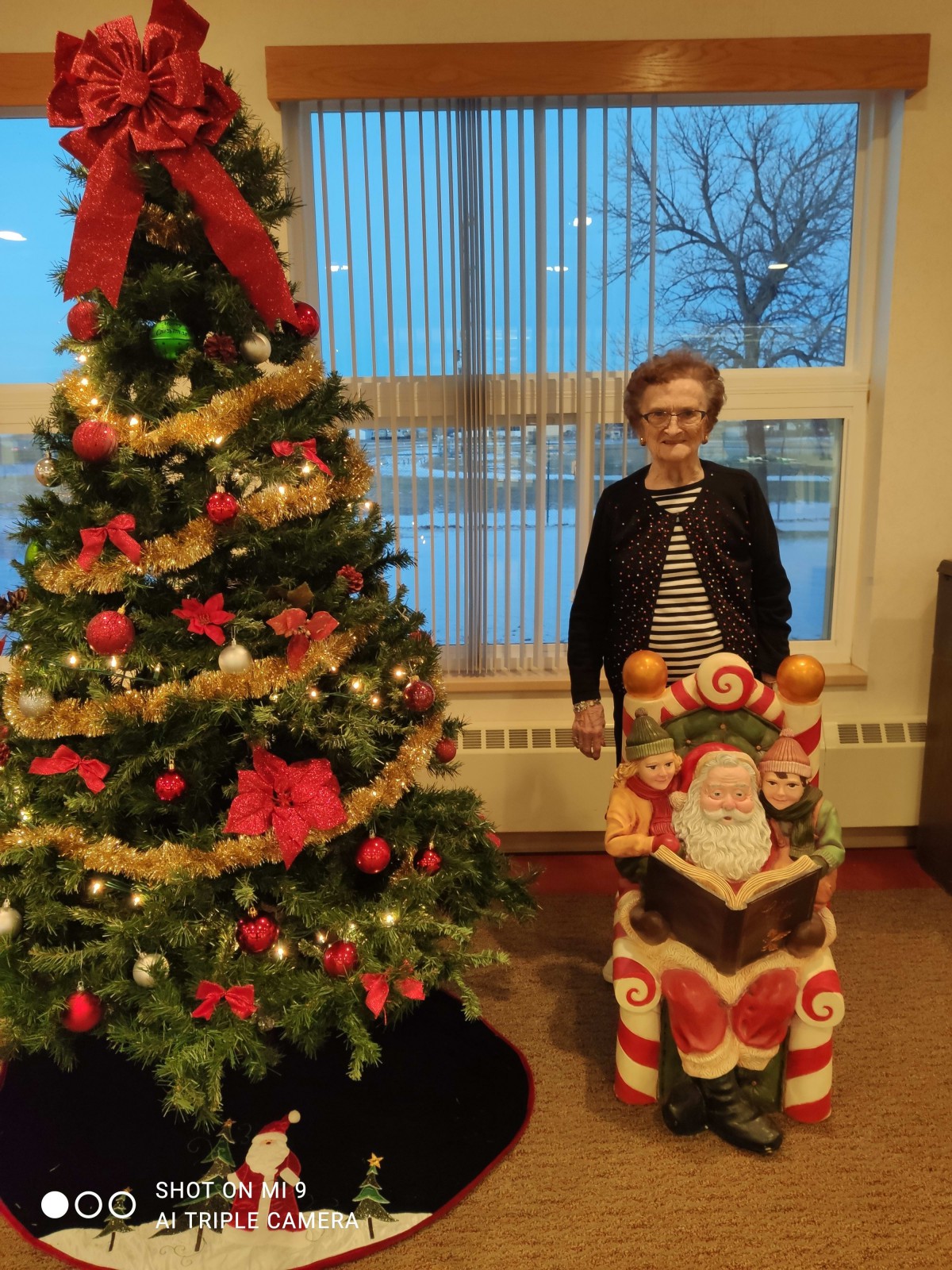 A person standing beside a decorated Christmas tree and a Santa figure indoors.