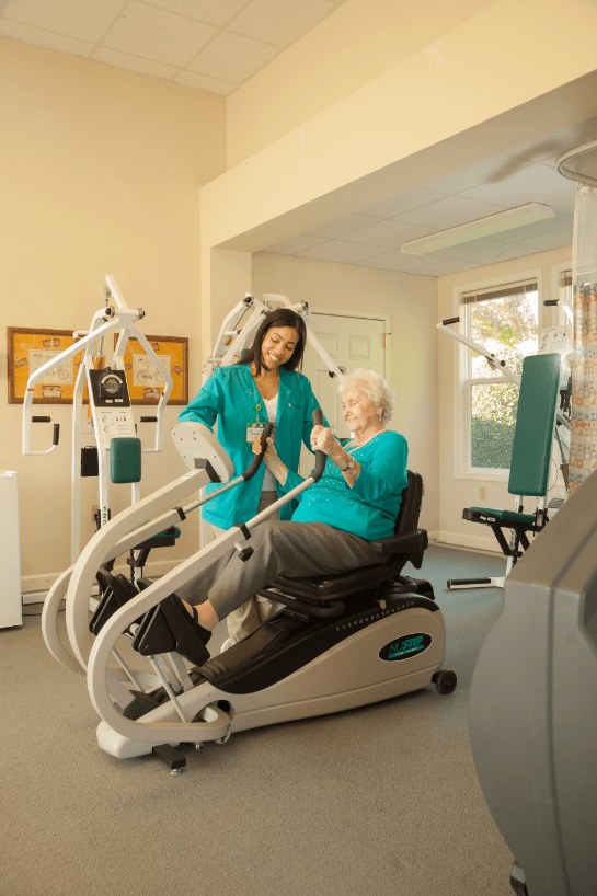 An elderly person receiving assistance while exercising on a fitness machine in a gym setting.