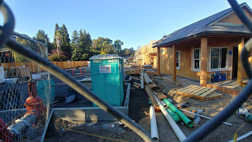 A view of a construction site featuring unfinished buildings, pipework, and a portable toilet under clear skies.