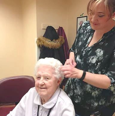 A caregiver styles the hair of an elderly woman indoors.