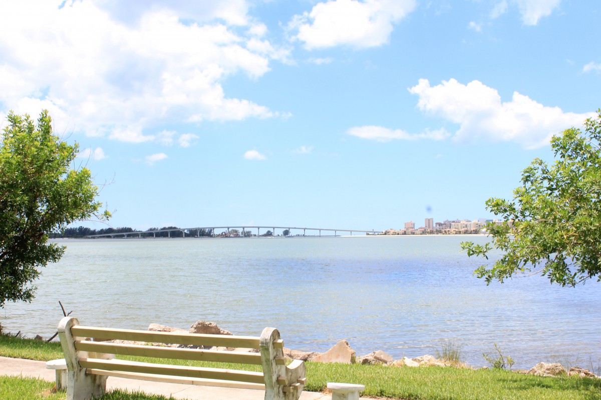 A serene view of a bench by the water with a bridge in the background under a bright, cloudy sky.