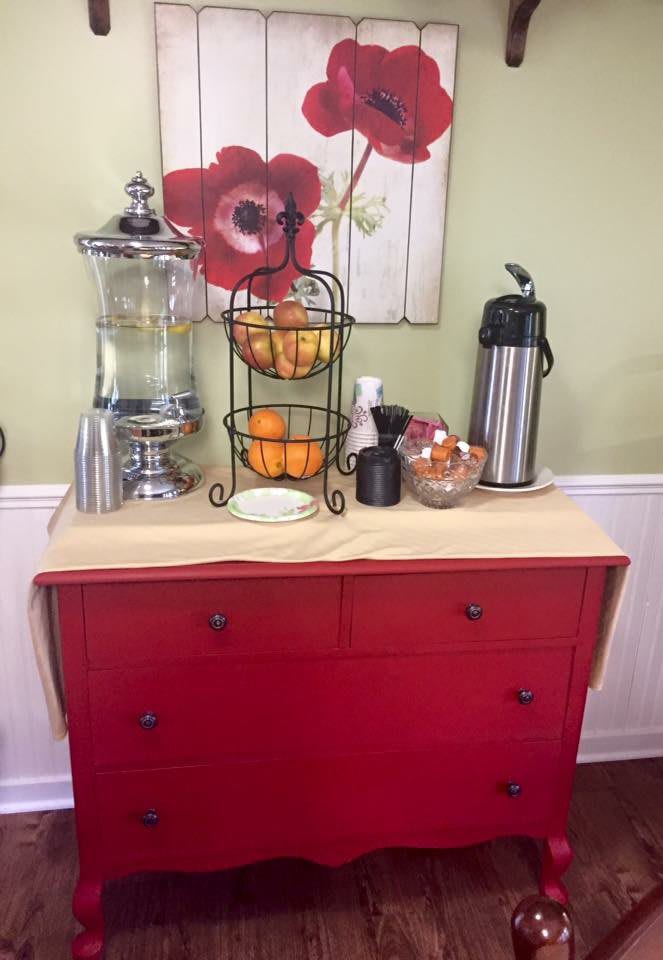 A neatly arranged beverage station with a water dispenser, coffee carafe, and a fruit basket on a red dresser.