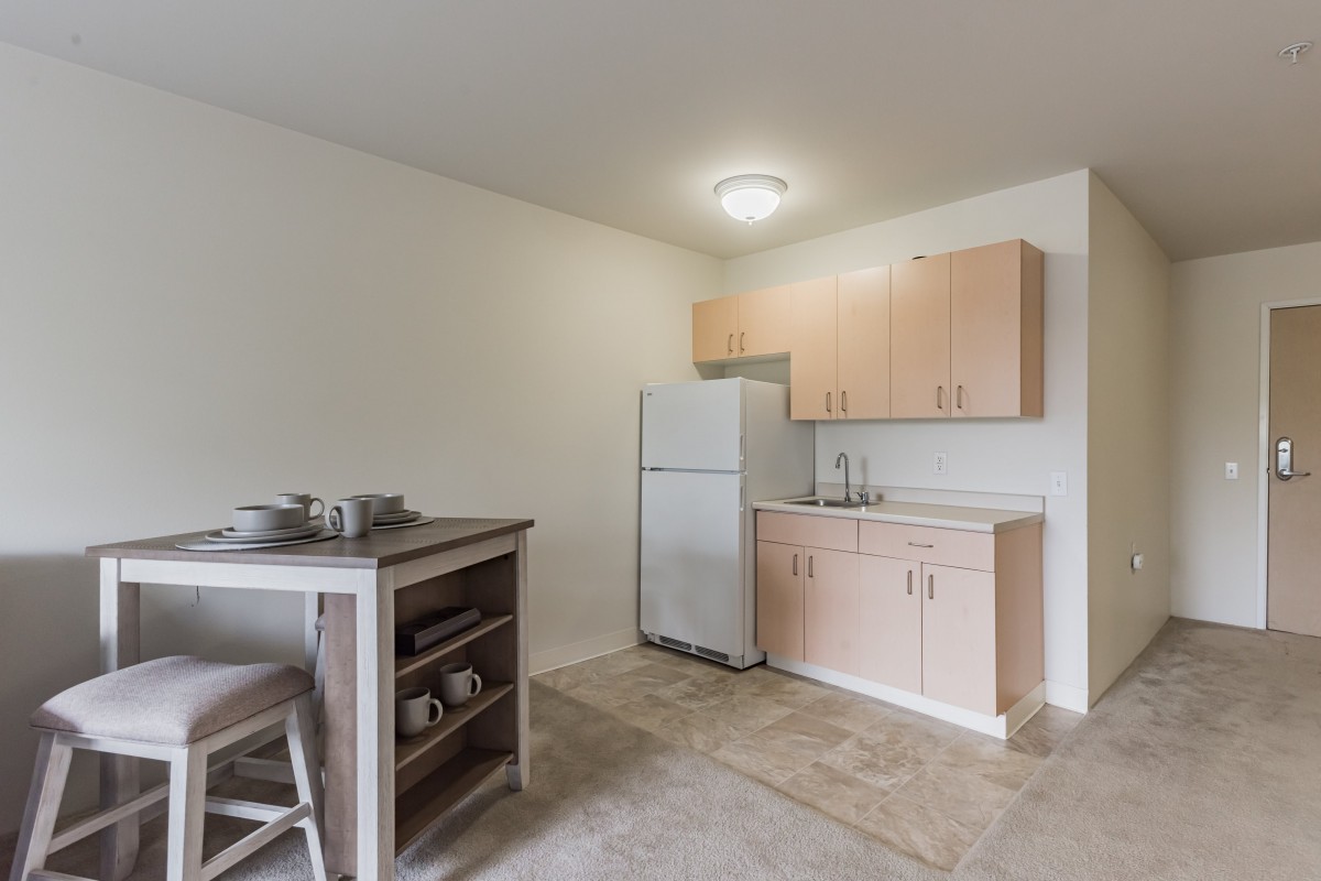 A minimalist kitchen with wooden cabinets, a white refrigerator, and a small dining table with stools.