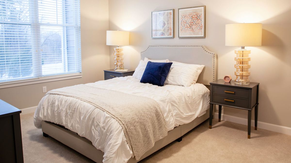 A cozy bedroom with a neatly made bed, featuring a navy blue accent pillow, two nightstands with lamps, and artwork on the walls.