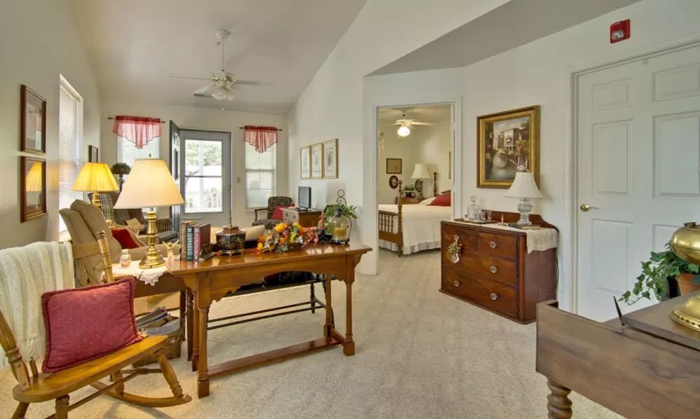 A cozy living room with a desk and traditional furnishings, offering a view into an adjacent bedroom.