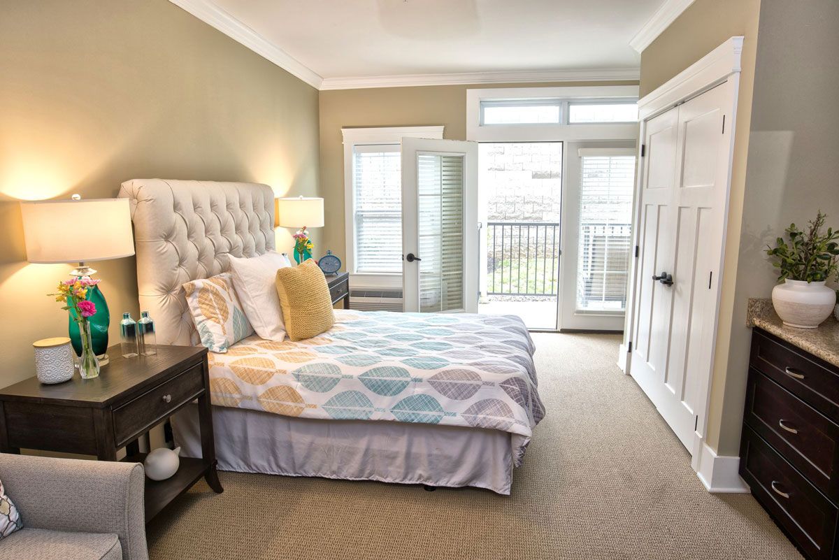 A cozy bedroom featuring a neatly made bed with colorful pillows, a bedside table with a lamp, and large windows letting in natural light.