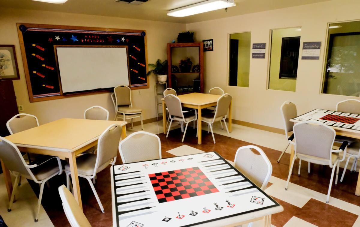 A classroom features several tables and chairs, a whiteboard, and a checkerboard game on one table.