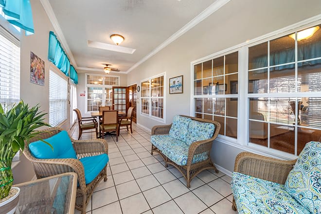 A bright and cozy sunroom with wicker furniture and a tiled floor, surrounded by large windows.