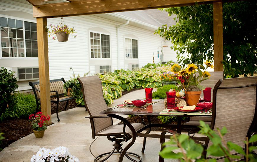 A cozy patio with a glass table and chairs, surrounded by lush garden foliage and vibrant flowers.