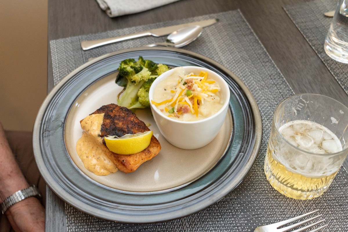 A served meal featuring grilled salmon with a lemon slice, a cup of soup topped with cheese and bacon bits, and steamed broccoli on a plate.