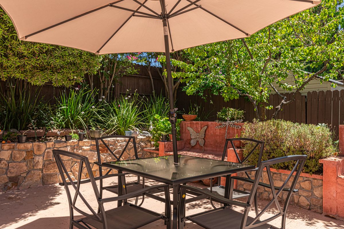 A sunny patio with a table, chairs, and a large umbrella surrounded by lush greenery.