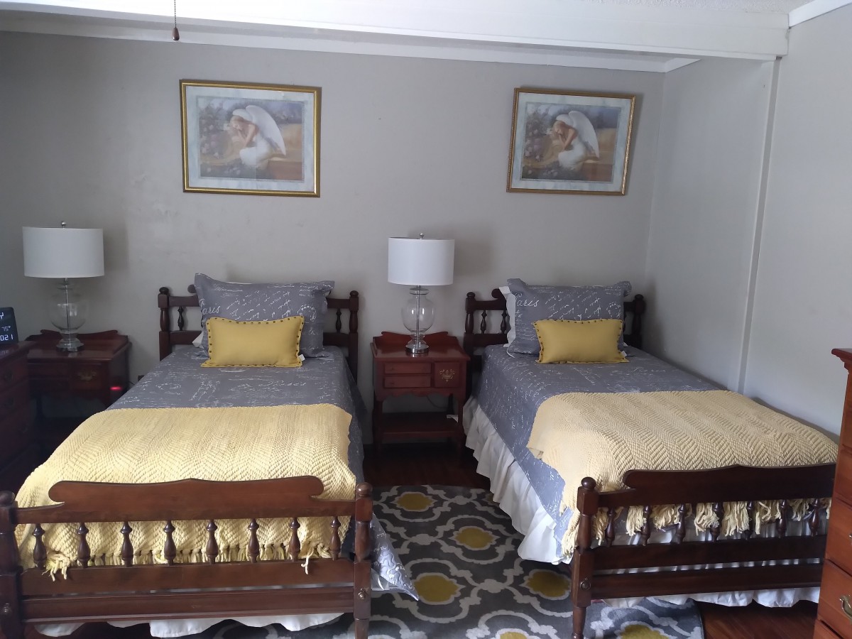 A cozy bedroom with twin beds featuring yellow and gray bedding, a central nightstand with a lamp, and decorative wall art above.