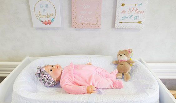 A baby in a pink outfit lies in a crib next to a stuffed bear, surrounded by nursery wall decorations.