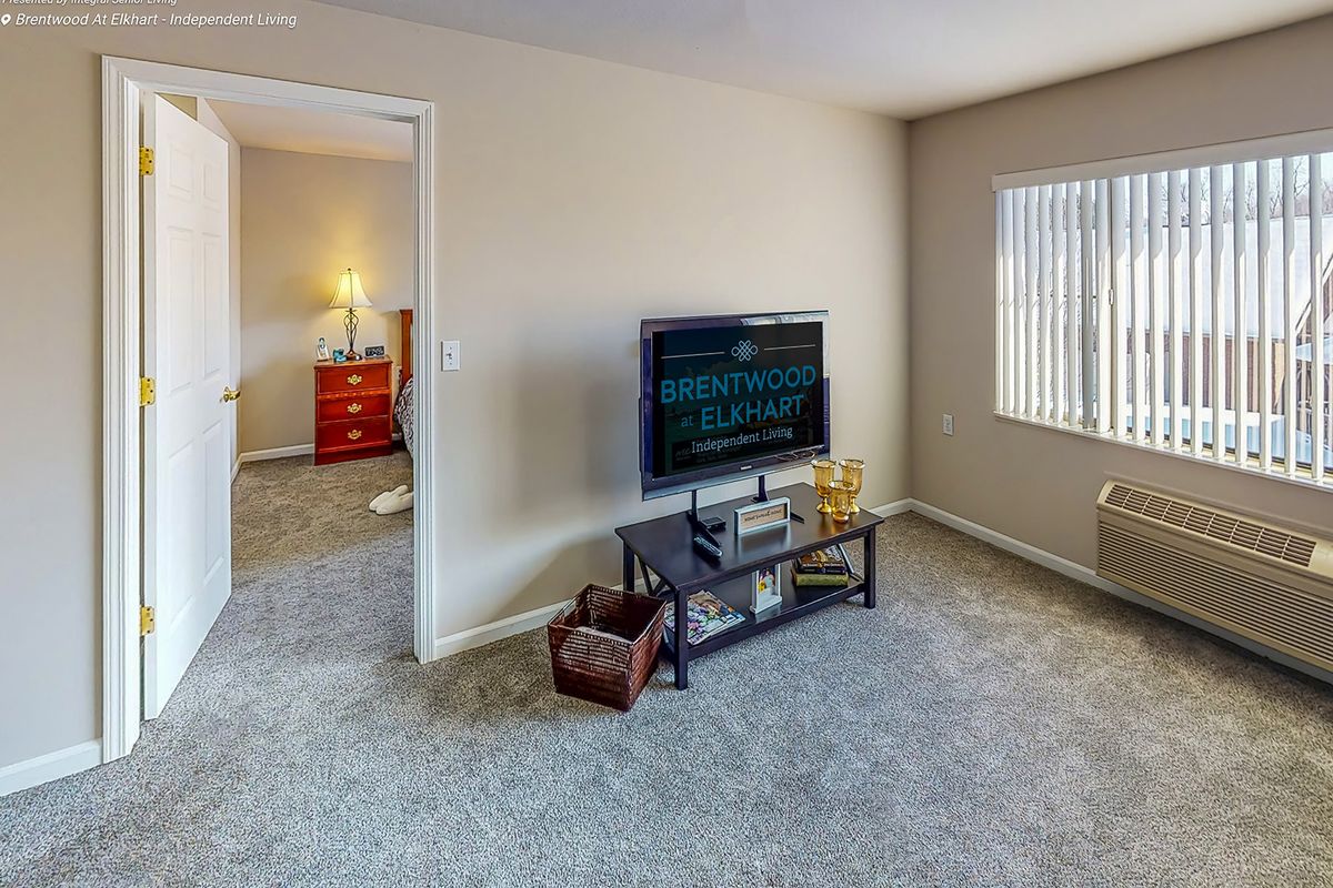 A cozy living room with a television, carpeted floor, and an adjacent bedroom visible through a doorway.