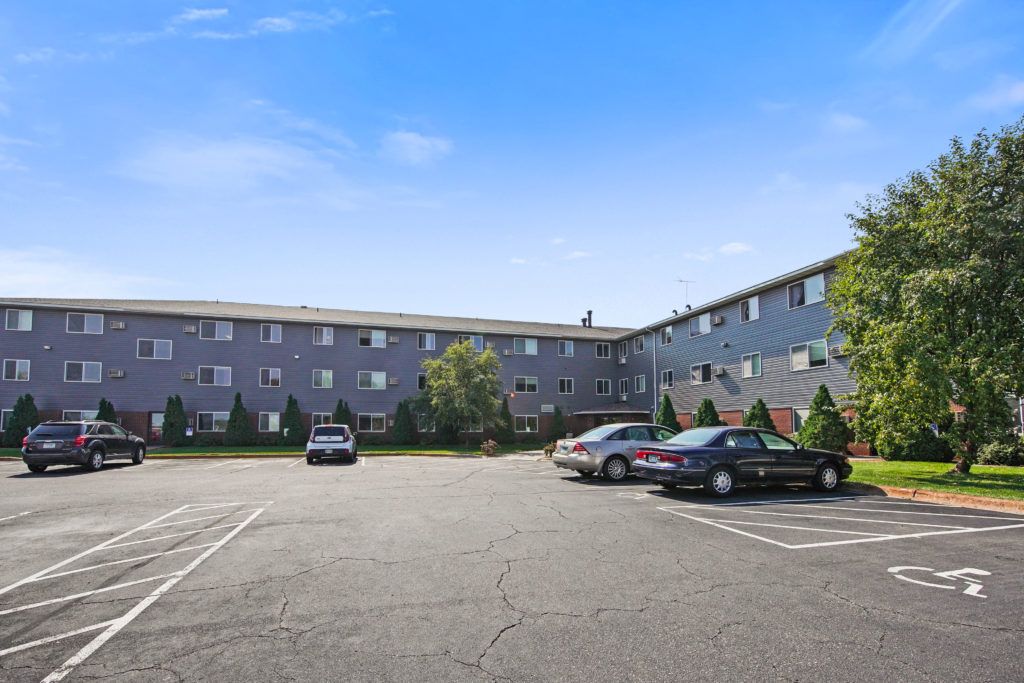An apartment complex with a parking lot in front, featuring several parked cars under a clear blue sky.