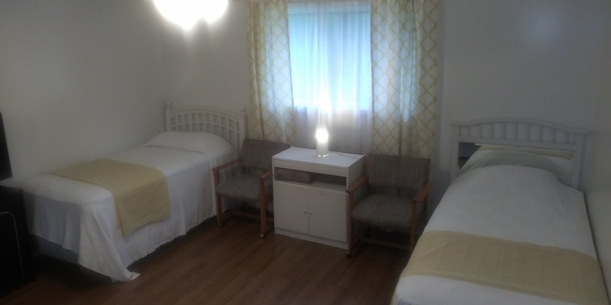 A simple bedroom with two beds, a nightstand with a lamp, and two chairs by the window.