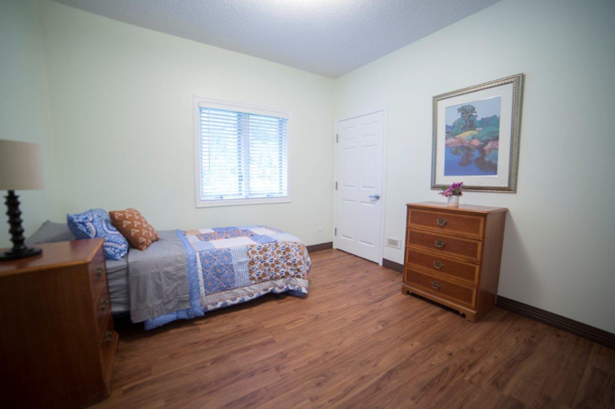 A minimalist bedroom features a single bed, wooden floors, and a dresser beneath a framed painting.