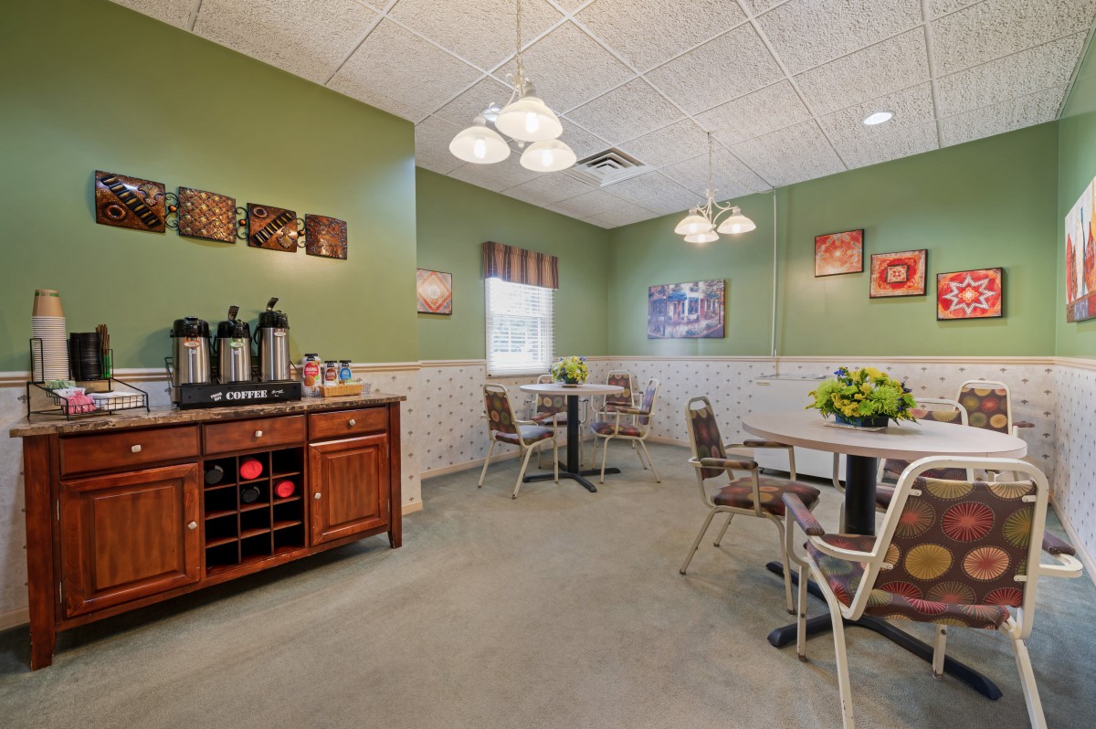 A cozy dining area with a coffee station, featuring tables and chairs under warm lighting, complemented by green walls and decorative art.