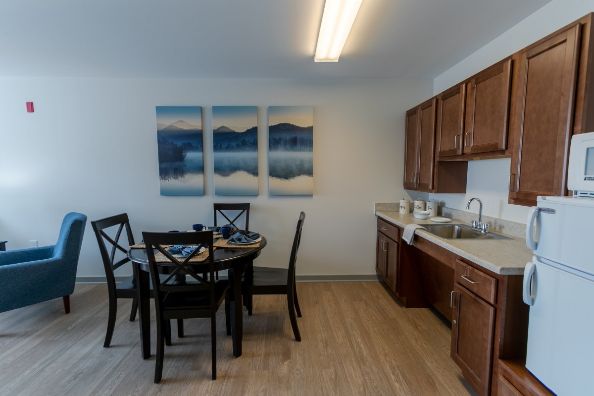 A modern dining area with a table and chairs, adjacent to a kitchen with wooden cabinets and a sink, featuring a triptych artwork on the wall.