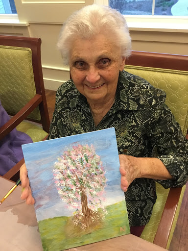 An elderly woman proudly displays her painting of a tree, showcasing her artistic talent and joy.