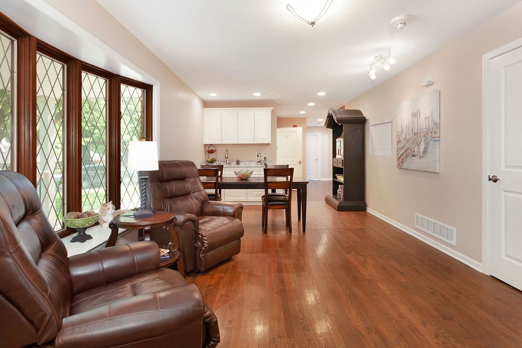 A spacious living room with hardwood flooring, featuring recliner chairs and an open layout leading to a modern kitchen and dining area.