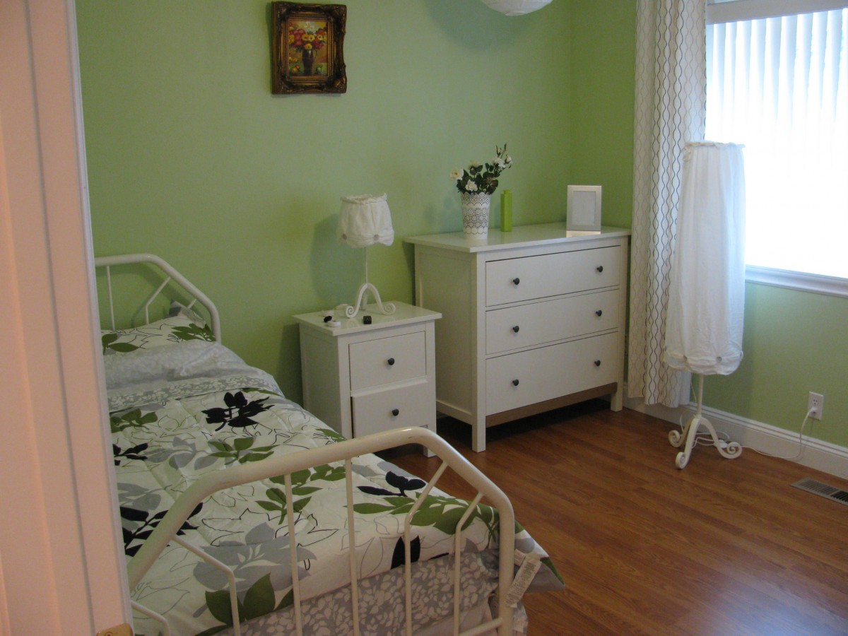 A cozy bedroom with green walls, featuring a single bed, a dresser, and a nightstand.