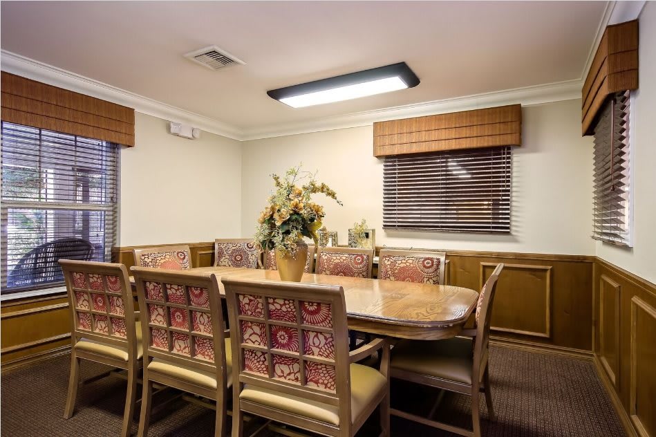 A cozy dining room with a large wooden table surrounded by upholstered chairs, adorned with decorative accents and window shades.
